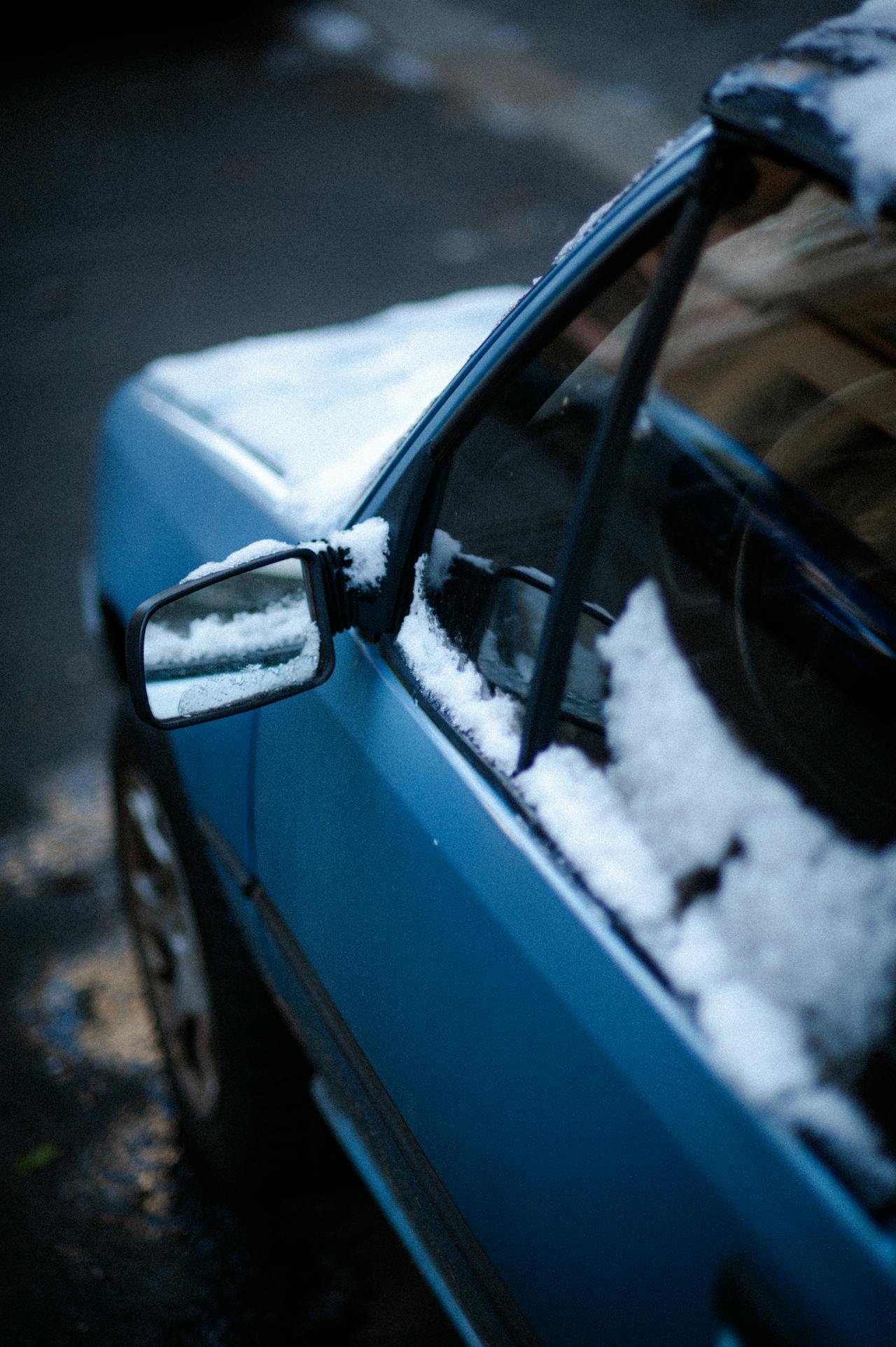 A blue car is shown with patches of snow on its side mirror, window, and roof.