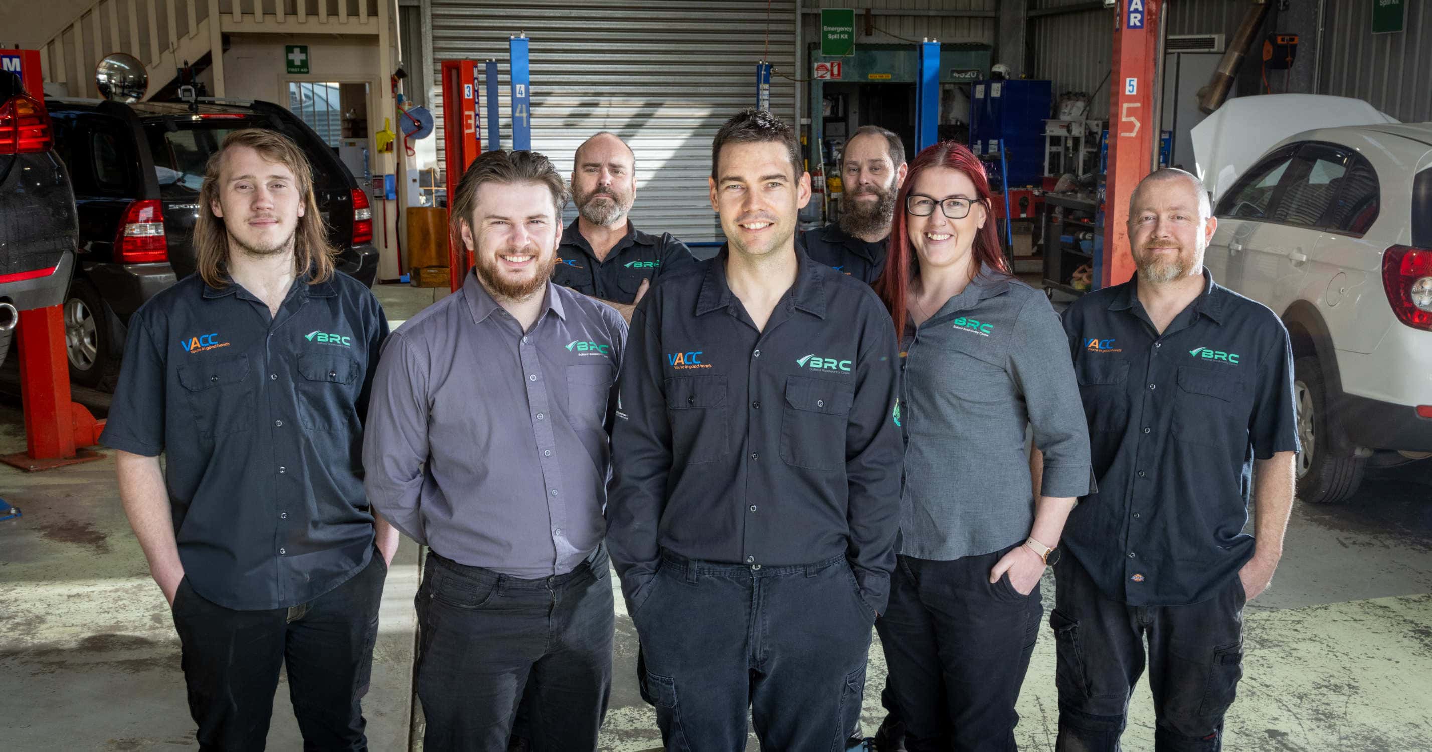 Group of seven auto mechanics in uniform standing inside a car repair shop on Ballarat Roadworthy, with car lifts and various vehicles in the background.